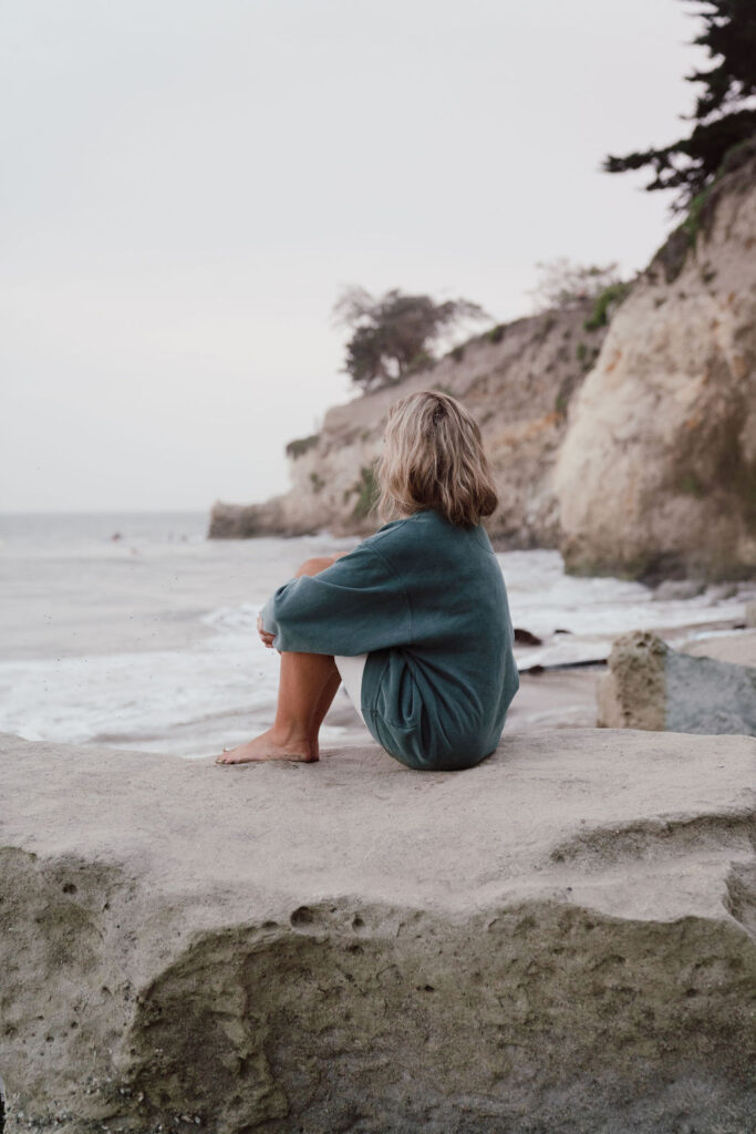 This image shows a woman sitting on the beach, looking out at the water. This episode explores how to untangle your emotions.