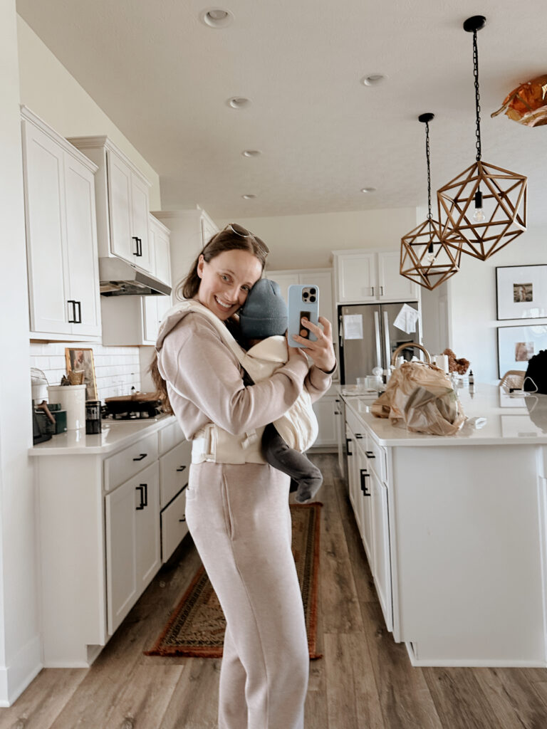 In this image, Jordan is wearing a sweatsuit, with her son in a baby carrier. This episode talks about recovery and movement postpartum.